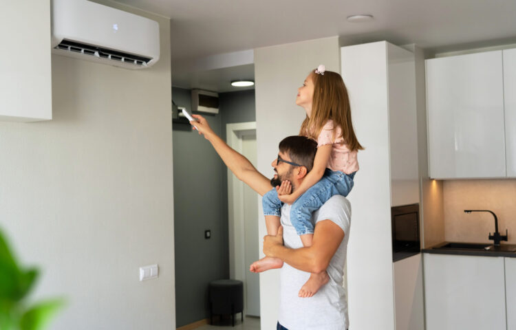Un père souriant porte sa fille sur ses épaules pendant qu'elle allume un climatiseur mural à l’aide d’une télécommande dans une cuisine moderne.